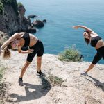 Two women stretching on a scenic cliff with the ocean view, embracing healthy outdoor living.