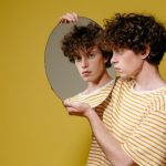 Studio portrait of a young man holding a mirror against a yellow background.
